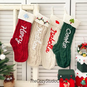 Peut inclure: Quatre bas de Noël en tricot torsadé rouge, crème et vert, chacun avec un revers blanc. Les bas sont personnalisés avec des noms brodés en blanc. L'un a un flocon de neige, un autre des lumières et un autre un sapin de Noël.