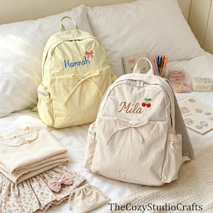 May include: A pale yellow backpack with the name Hannah and a pink bow embroidered on it, and a cream-coloured backpack with the name Mila and a red cherry motif embroidered on it. Both bags feature a front pocket with a decorative tie detail.