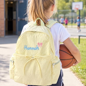 May include: A pale yellow backpack featuring a top handle, two side pockets with zips, and a front pocket with a decorative drawstring bow. The name Hannah is embroidered in blue script with a small pink bow icon above the name on the front.