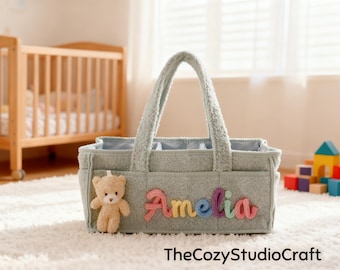May include: A gray baby diaper caddy with a teddy bear and the name "Amelia" in colorful letters. The caddy has two handles and several pockets. A wooden crib and toys are in the background.