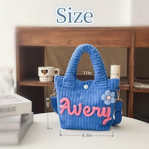 May include: Blue corduroy tote bag with the name "Avery" in pink script. The bag has a flower accent and a shoulder strap. It measures 11 inches wide and 8.3 inches high.