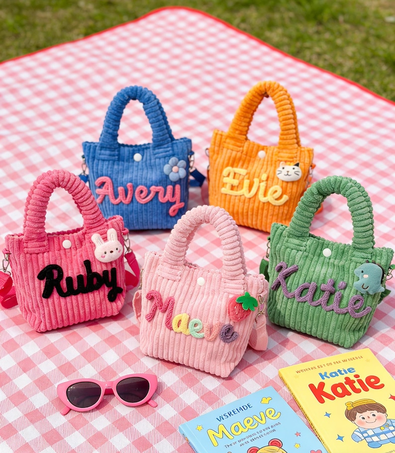 May include: Five colorful corduroy tote bags with names embroidered on them. The bags are in shades of blue, orange, pink, light pink, and green. Each bag has a matching handle and a small decorative charm. Pink sunglasses and two books are in the foreground.