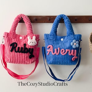 May include: Two corduroy tote bags, one pink and one blue, each with a name embroidered on the front. The pink bag has the name "Ruby" in black and a white bunny. The blue bag has the name "Avery" in pink and a blue flower.