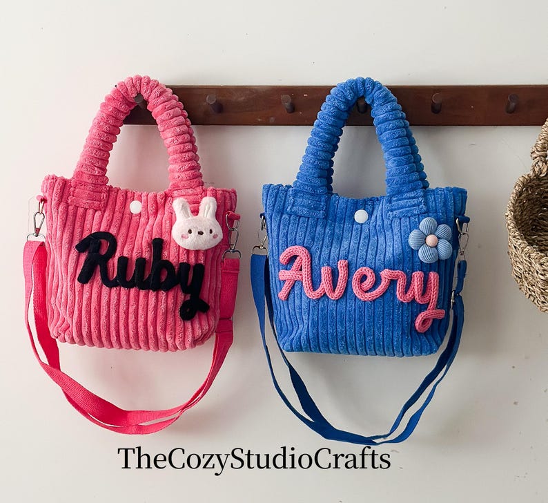 May include: Two corduroy tote bags, one pink and one blue, each with a name embroidered on the front. The pink bag has the name "Ruby" in black and a white bunny. The blue bag has the name "Avery" in pink and a blue flower.
