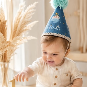 Könnte beinhalten: Ein blauer, kegelförmiger Partyhut mit einem hellblauen Wollbommel an der Spitze, einer weißen Spitzenborte am unteren Rand und gestickten Details wie einem kleinen Kuchen, Sternen und dem Namen Noah in weißem Garn mit einem dünnen Gummiband.