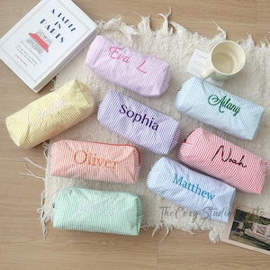 May include: A selection of personalised pencil cases in pastel shades, such as pink, yellow, lilac, orange, green, blue, and red. Each case has a different name embroidered in a contrasting colour. The cases are made of seersucker fabric.