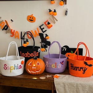 Peut inclure: Plusieurs articles sur le thème d'Halloween, dont quatre paniers tressés blancs, noirs, violets et oranges, chacun avec un nom brodé sur le côté. Une citrouille sculptée avec un intérieur éclairé est devant les paniers. Décorations d'Halloween en arrière-plan.