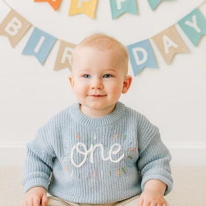May include: A light blue long-sleeve knit jumper featuring the word one embroidered in white script on the chest, surrounded by small, colourful vertical stitch accents. The jumper has a ribbed crew neckline and ribbed cuffs.