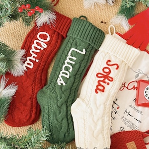 May include: Three cable-knit Christmas stockings in red, green, and cream, each with a name embroidered in a contrasting colour: Olivia, Lucas, and Sofia. The stockings are decorated with a cable knit pattern and hang from loops. Festive greenery and berries surround the stockings.