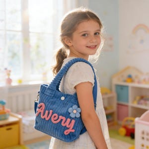 May include: A blue corduroy tote bag with the name "Avery" in pink script. The bag has a blue flower applique and a shoulder strap. The bag is being carried by a young person.