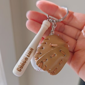 Peut inclure: Porte-clés sur le thème du baseball avec une mini-batte de baseball portant l'inscription "COACH LIAM", un gant de baseball et une balle de baseball. Le porte-clés est argenté. Le gant de baseball est marron et la balle de baseball est blanche avec des coutures rouges.