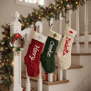 May include: Three cable-knit Christmas stockings hanging from a staircase railing. The red stocking is embroidered with the name Henry, the green with Cookie, and the cream with Isabella. Each features a ribbed cuff with a small festive icon.