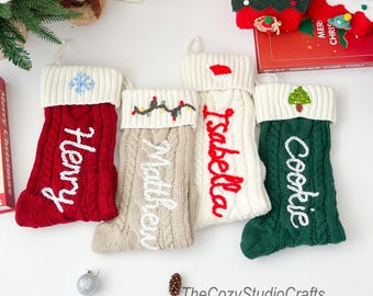 May include: A collection of personalized Christmas stockings in red, beige, white, and green. Each stocking features a cable knit design with a white cuff and a name embroidered in a contrasting color. Festive decorations include a snowflake, lights, a Santa hat, and a Christmas tree.