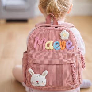 May include: A pink corduroy backpack with the name "Maeve" in colourful knitted letters. Features a white flower and a bunny applique. Includes side pockets and tassel details. The Cozy Studio Crafts logo is at the bottom.