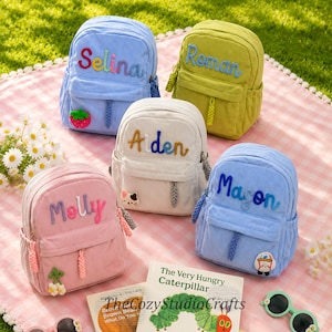Peut inclure: Cinq sacs à dos en velours côtelé bleu clair, vert citron, crème, rose et bleu ciel. Chaque sac présente un prénom brodé en écriture colorée, une poche avant zippée avec une tirette tressée et un petit écusson décoratif.