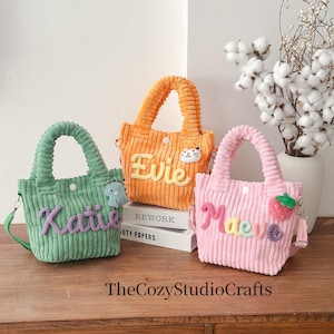 May include: Three corduroy tote bags in green, orange, and pink, each with a name embroidered on the front. The green bag has a blue dinosaur, the orange bag has a cat, and the pink bag has a strawberry. The bags have handles and shoulder straps.