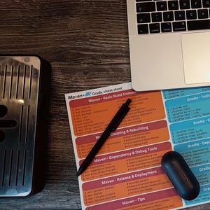 May include: A close-up of a printed cheat sheet with instructions for Maven and Gradle, two popular build automation tools. The sheet is divided into sections with headings like "Basic Build Commands", "Cleaning & Rebuilding", "Dependency & Debug Tools", "Testing & Profiling", and "Publishing & Release". A black pen and a dark blue wireless earbud are also visible on the sheet.