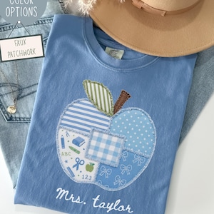 May include: A blue t-shirt featuring a large apple-shaped appliqué made of various blue and white patterned fabric patches. The apple design includes school-themed icons like pencils, books, and numbers. The text "Mrs. Taylor" is printed in white below the apple.