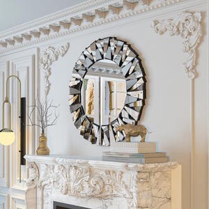 Peut inclure: Un miroir rond décoratif avec un cadre noir et une mosaïque de morceaux de verre réfléchissants. Le miroir est suspendu au-dessus d'une cheminée blanche ornée. Une figurine de rhinocéros dorée et des livres sont sur le manteau.