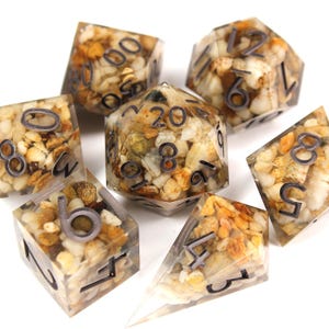 May include: Set of seven polyhedral dice with a clear resin base and embedded small stones. The dice are numbered 1-20, 4, 6, 8, 10, and 12. The dice are a light brown colour with a speckled appearance.