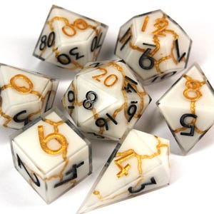 May include: Set of seven white dice with black and gold numbers and a cracked, marbled effect. The dice are for role-playing games.