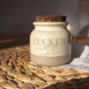 Könnte beinhalten: Eine Keramik-Zuckerdose mit einem Korkverschluss. Die Dose ist beige mit einem braunen Band um den Boden. Der Text "ZUCKER mit Löffel Glas in jeder Tasse" ist auf die Dose gedruckt.