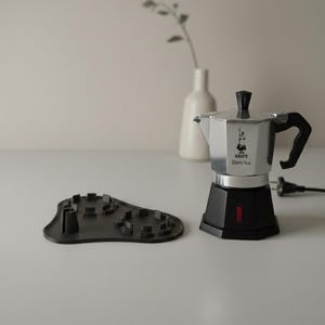 Op de afbeelding: Een zilveren en zwarte elektrische koffiezetter met de tekst "Bialetti Elettrika" op de voorkant. Een zwarte, getextureerde onderzetter staat links. Een witte vaas met een takje groen staat op de achtergrond.