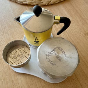 Op de afbeelding: Een gele en zilveren espressomaker voor op het fornuis met een zwarte handgreep en knop, samen met de onderdelen. De maker heeft het Bialetti-logo en tekst. De onderdelen omvatten een filtermand en een deksel met de tekst "MADE BY BIALETTI SINCE 1933 ITALIAN QUALITY & DESIGN BS065D."