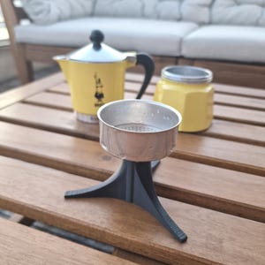 Könnte beinhalten: Ein gelber Espressokocher mit schwarzem Griff und Deckel, ein silberner Filter und ein gelber Behälter mit silbernem Deckel stehen auf einem Holztisch. Der Espressokocher hat ein schwarzes Logo.