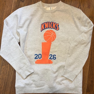 May include: A light gray crewneck sweatshirt featuring a graphic print on the chest. The design includes the word KNICKS in blue letters above an orange basketball trophy icon, with the numbers 20 and 26 printed in blue on either side of the base.