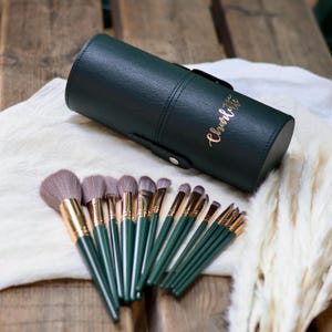 Könnte beinhalten: Ein Set Make-up-Pinsel mit dunkelgrünen Griffen und goldenen Zwingen, angeordnet mit einem passenden dunkelgrünen zylindrischen Etui. Das Etui hat einen Griff und den Namen "Charlotte" in goldener Schrift. Die Pinsel haben weiche, hellfarbige Borsten.