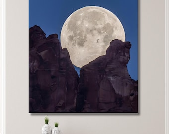 May include: A large canvas print depicts a silhouetted figure walking a tightrope against a backdrop of a full moon and dark rock formations. The moon is bright white against a deep blue sky, creating a dramatic contrast. The artwork is displayed on a wall in a well-lit room.