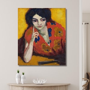 Puede incluir: Una vibrante pintura al óleo de una mujer con cabello y ojos oscuros, vestida con una prenda roja con patrones florales amarillos y azules. El fondo es de un amarillo brillante, y un abanico negro descansa sobre la mesa. La obra de arte se exhibe en una pared.