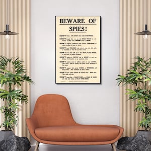 May include: A framed vintage poster with the text "BEWARE OF SPIES!" in bold letters. The poster features a list of instructions in black text on a cream-colored background, warning against sharing information. The poster is displayed in a living room setting.