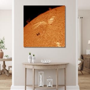 May include: A canvas print of the sun's surface, showcasing intricate patterns and textures in shades of orange and yellow. A small, dark shape, possibly a spacecraft, is visible against the solar backdrop. The artwork is displayed on a white wall.