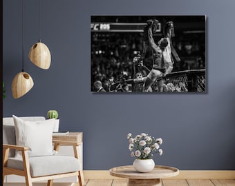 May include: Black and white photograph of a mixed martial arts fighter celebrating a victory, holding championship belts above his head. The image is from UFC 205, with a crowd in the background.