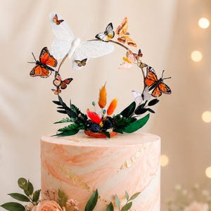 Puede incluir: Pastel de dos pisos con glaseado veteado melocotón y blanco, adornado con detalles de pan de oro y flores frescas. Un adorno para pastel con tema de mariposas presenta un anillo de mariposas. El pastel está sobre un soporte dorado.