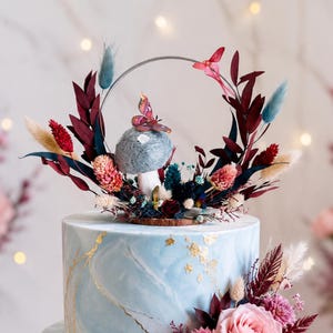 Puede incluir: Tarta de dos pisos con glaseado azul claro con efecto mármol y detalles dorados. La tarta está decorada con flores rosas y burdeos, y un adorno circular con un diseño de mariposa y hongo. El texto "kirahley kreations" es visible.