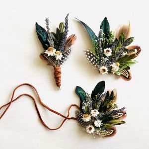 Op de afbeelding: Drie handgemaakte bloemen- en verenaccessoires. De stukken hebben een mix van donkergroene, bruine en gespikkelde veren, gedroogde witte bloemen en lavendeltakjes. Eén stuk is een corsage gewikkeld in bruin leer, en een ander is een hoofdband met een bruin leren koord.