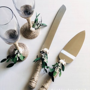 May include: Two champagne flutes and a cake knife and server set with rustic twine wrapped handles and greenery accents. The twine is wrapped around the base of the flutes and the handles of the cake cutting tools.  The greenery is a mix of small white flowers and green leaves.