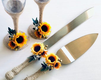 May include: A wedding cake cutting set with two champagne flutes decorated with sunflowers and twine. The knife and server handles are also wrapped in twine and decorated with sunflowers.