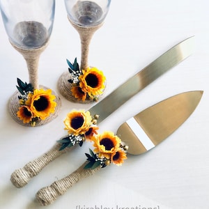 May include: A wedding cake cutting set with two champagne flutes decorated with sunflowers and twine. The knife and server handles are also wrapped in twine and decorated with sunflowers.