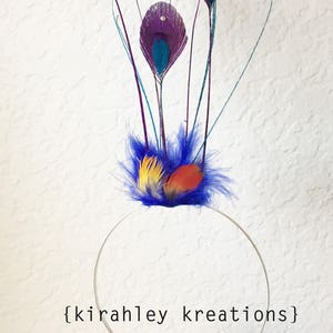 May include: A handmade headband featuring peacock feather accents. The headpiece has a silver band with a cluster of purple, teal, yellow, and orange feathers. The text "{kirahley kreations}" is printed on the image.