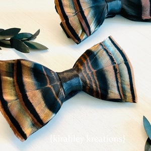 May include: Two bow ties made from feathers, featuring a pattern of brown, black, and tan. The bow ties are displayed on a white surface, with dark green leaves in the background. The text "{kirahley kreations}" is visible.