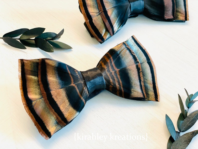May include: Two bow ties made from feathers, featuring a pattern of brown, black, and tan. The bow ties are displayed on a white surface, with dark green leaves in the background. The text "{kirahley kreations}" is visible.