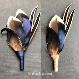 May include: Two feather boutonnieres. One is wrapped with navy blue ribbon, the other with twine. The feathers are a mix of black, brown, white, and blue. The boutonnieres are set against a gray background.