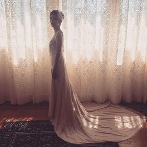 May include: A woman in a white wedding dress with a long train stands in front of a lace curtain. The dress has a fitted bodice and a flowing skirt. The woman is looking to the side.