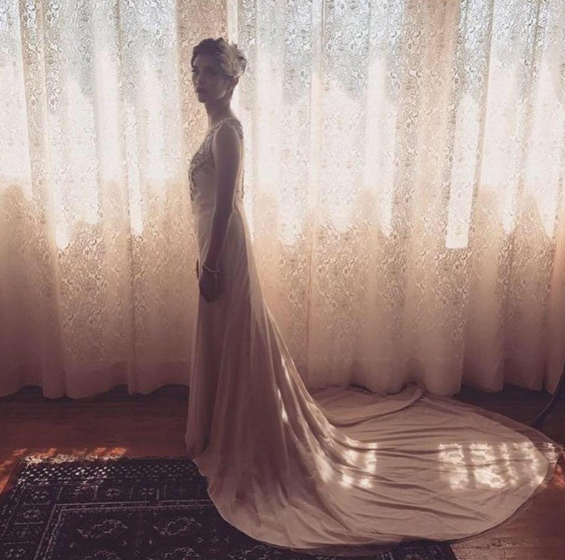 May include: A woman in a white wedding dress with a long train stands in front of a lace curtain. The dress has a fitted bodice and a flowing skirt. The woman is looking to the side.