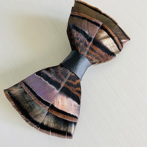 May include: A handmade feather bow tie with a black leather band. The bow tie features brown, black, and purple feathers. The text "{kirahley kreations}" is at the bottom of the image.