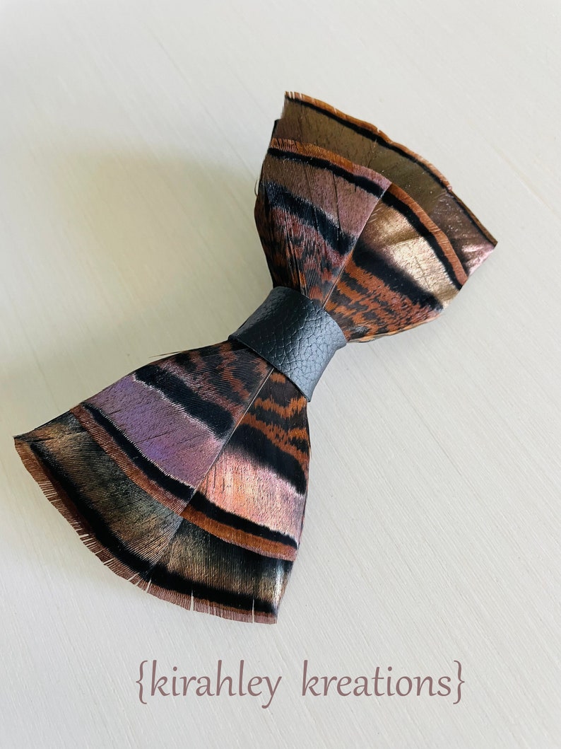 May include: A handmade feather bow tie with a black leather band. The bow tie features brown, black, and purple feathers. The text "{kirahley kreations}" is at the bottom of the image.